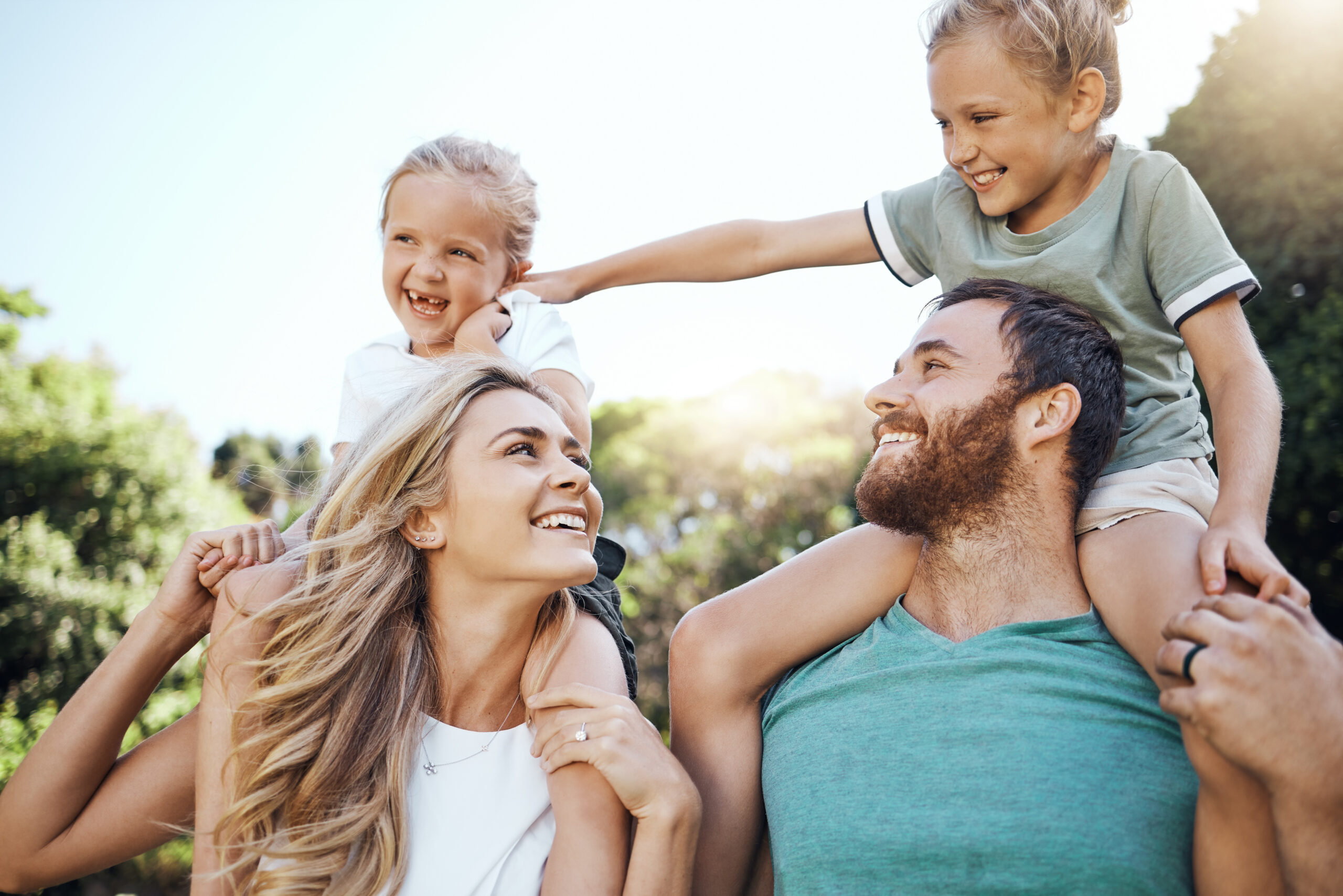 Mom and dad with kids on their shoulder in the garden, playing together. Love, young and happy family bond and have fun in a park on a summer day. Father, mother and daughters enjoy being in nature
